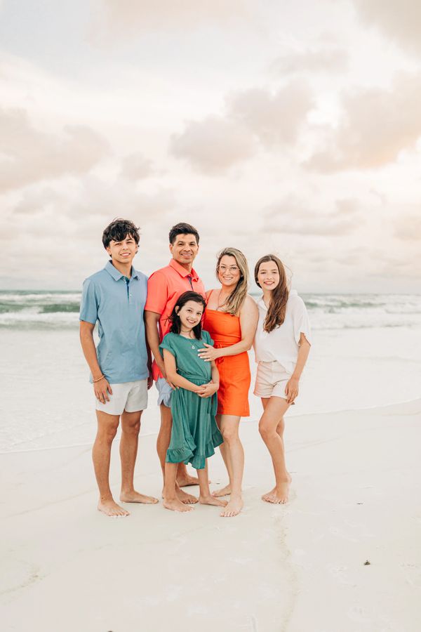 A happy family of five posing barefoot on a beach at sunset.