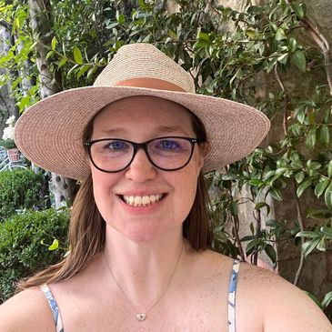 A smiling woman wearing glasses and a straw hat outdoors, surrounded by greenery.