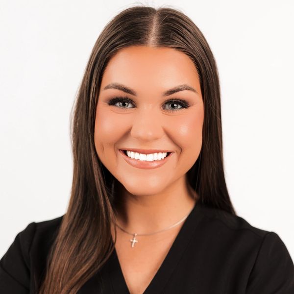 Smiling woman with long brown hair wearing a black top and cross necklace.