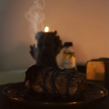 Smoldering sage bundle releasing smoke on a plate in a dimly lit setting.