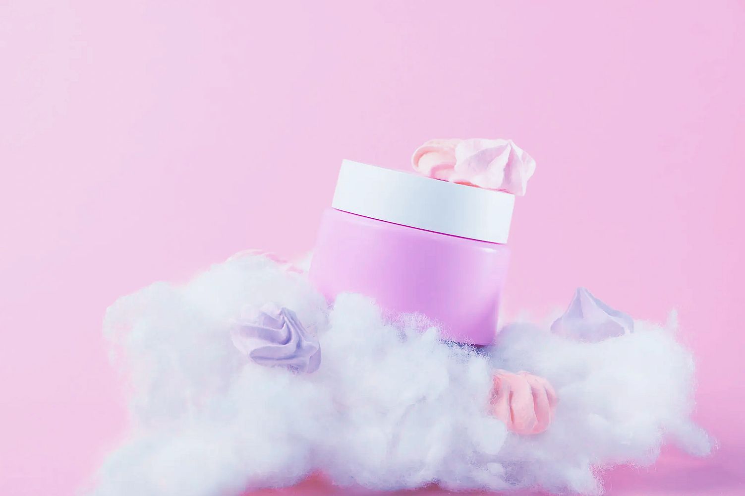 Pink cosmetic jar surrounded by fluffy white cotton and pastel meringue decorations.