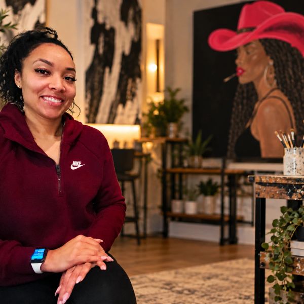Woman smiling in casual wear indoors with vibrant painting behind.