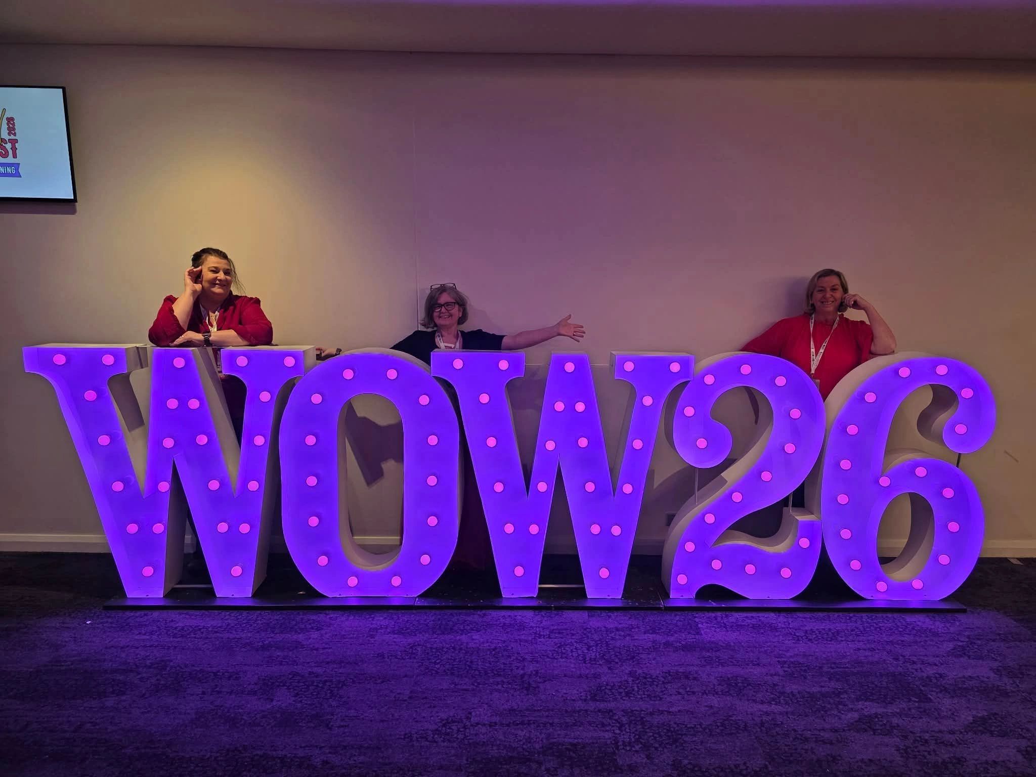 Three women posing behind illuminated letters spelling WOW26.