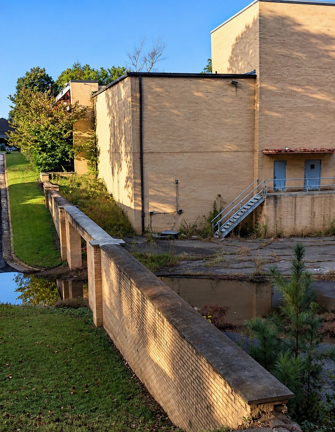 Sunlit brick building with a staircase and overgrown area.