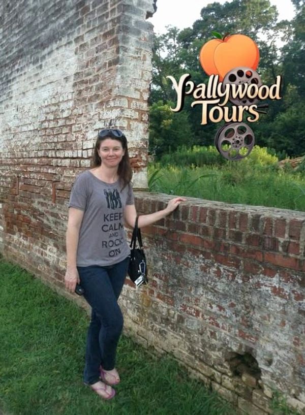 Woman standing by an old brick wall with Y'allywood Tours sign.