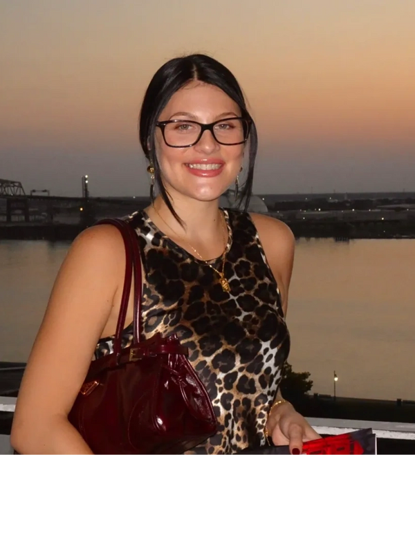 Smiling woman in glasses with a leopard print top at sunset by a river and bridge.