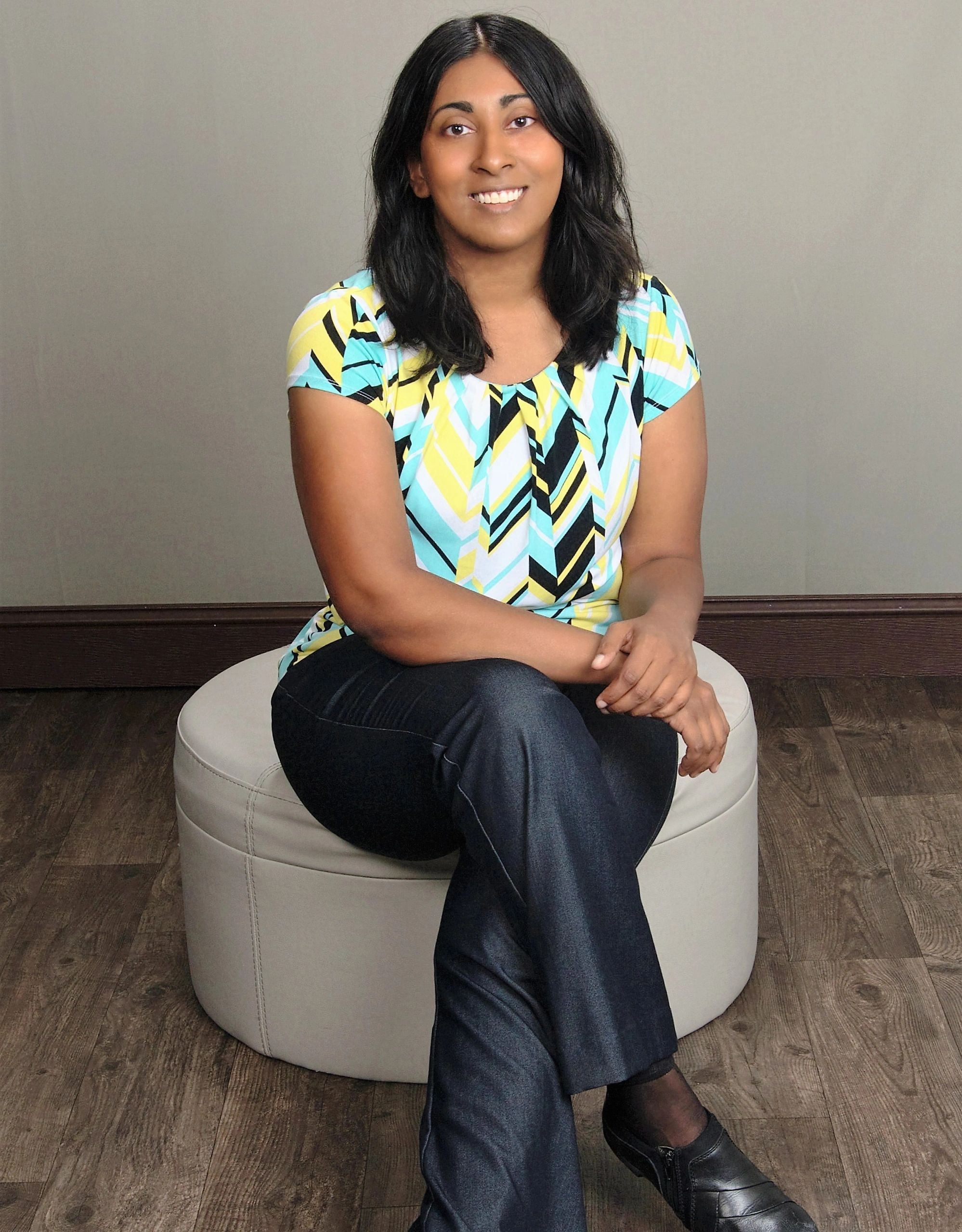 Smiling woman sitting on a white round ottoman against a plain wall.