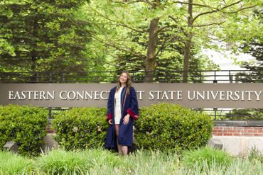 A graduate in cap and gown standing by the Eastern Connecticut State University sign.