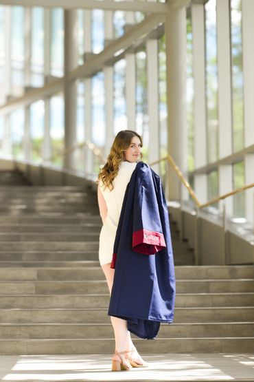 Woman holding a graduation gown on stairs in a sunlit building.