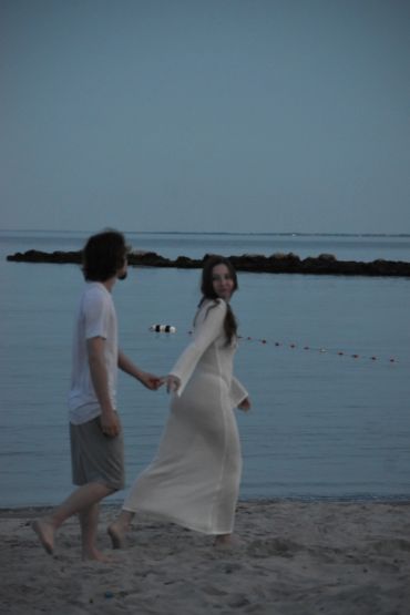 A couple holding hands and walking barefoot on a beach near the water.