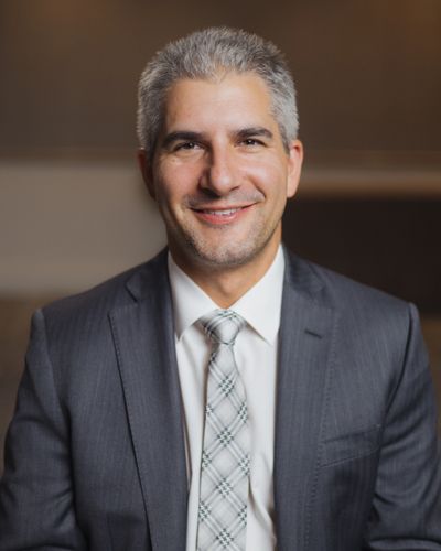 Smiling man in a gray suit and patterned tie against a blurred background.