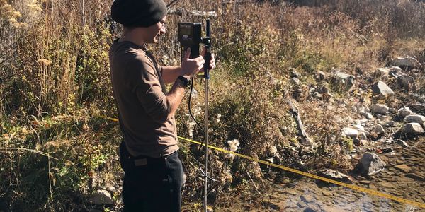 Person using a measuring device by a stream in a natural setting.