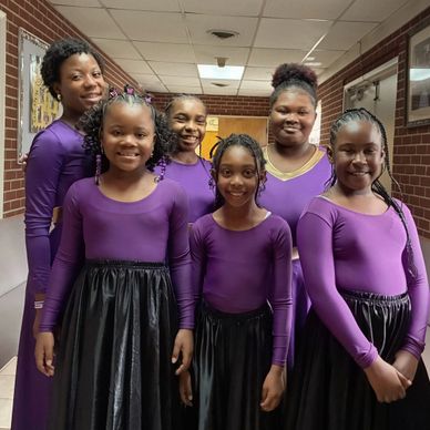 Six girls in purple tops and black skirts smiling indoors.
