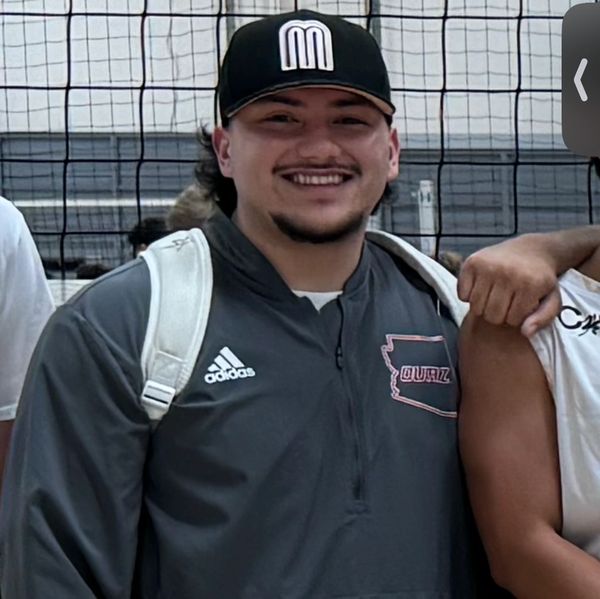 A smiling man in a black cap and gray Adidas jacket stands in front of a volleyball net.