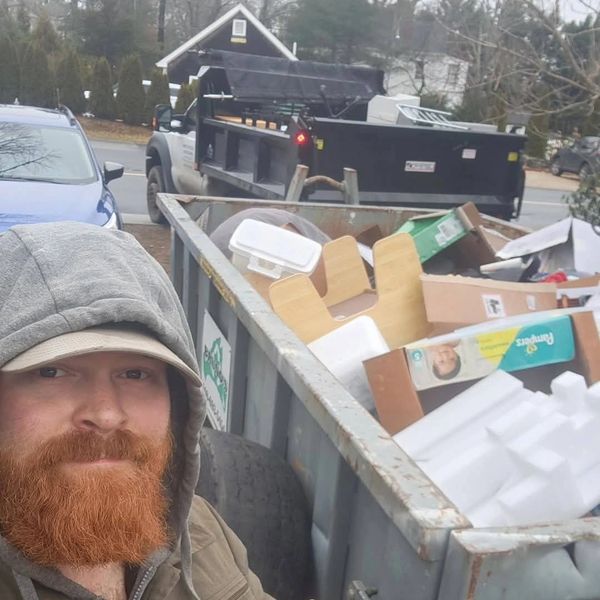 Man with red beard in hoodie takes selfie near a dumpster filled with trash and cardboard boxes.