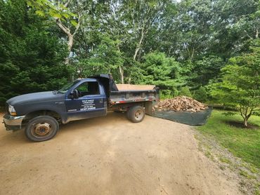 A blue dump truck unloading a pile of dirt in a wooded area.