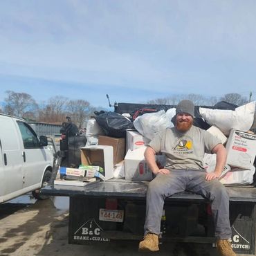 Man with a beard sitting on a truck bed surrounded by bags and boxes.