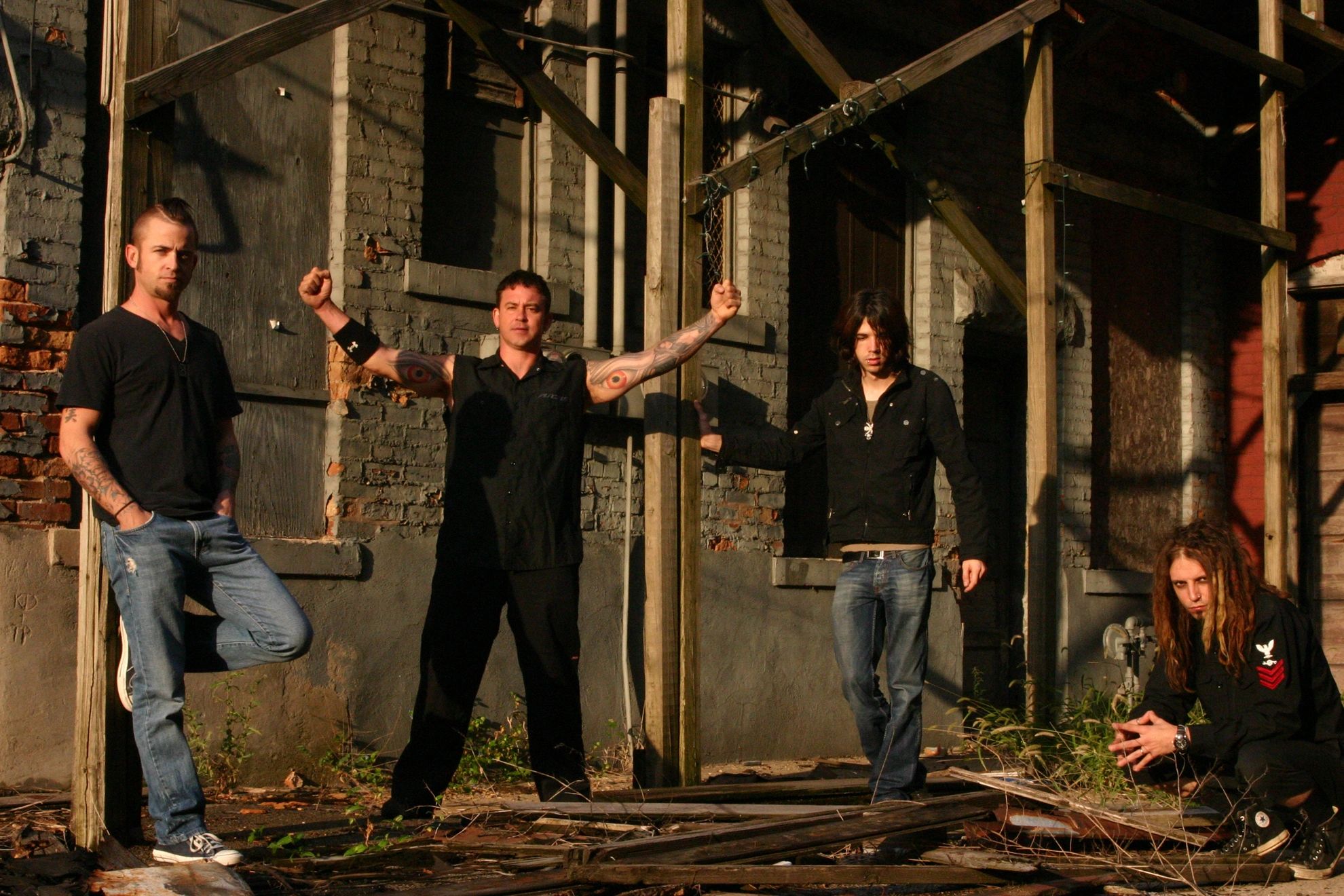 Four men posing in front of a weathered brick building with wooden supports.