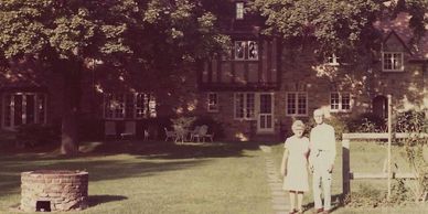 Elderly couple standing on a garden path in front of a large stone house.