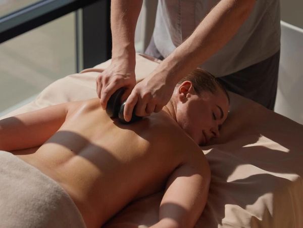 A woman receiving a back massage with a massage tool in a sunlit room.