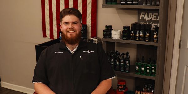 Barber smiling in front of grooming products and an American flag.