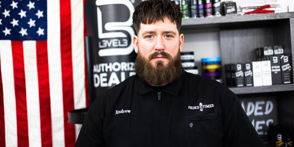 Bearded man in a black jacket stands in front of an American flag and shelves with products.