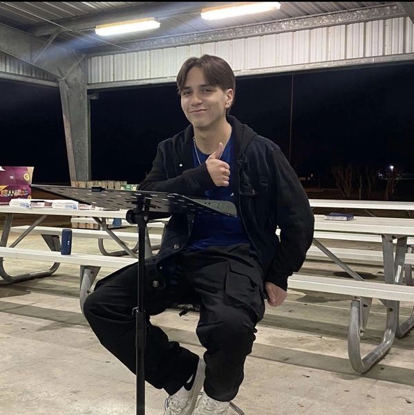 A young man giving a thumbs-up while sitting at a music stand in an outdoor pavilion.