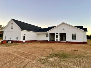 Modern white farmhouse with a black roof on a bare dirt lot at sunset.