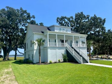 White two-story house with a front porch and stairs, surrounded by green grass and trees.