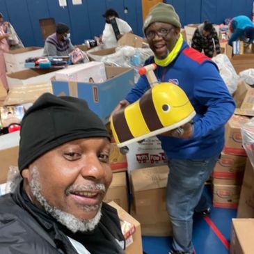 Two men smiling amidst boxes, one holding a bee toy.