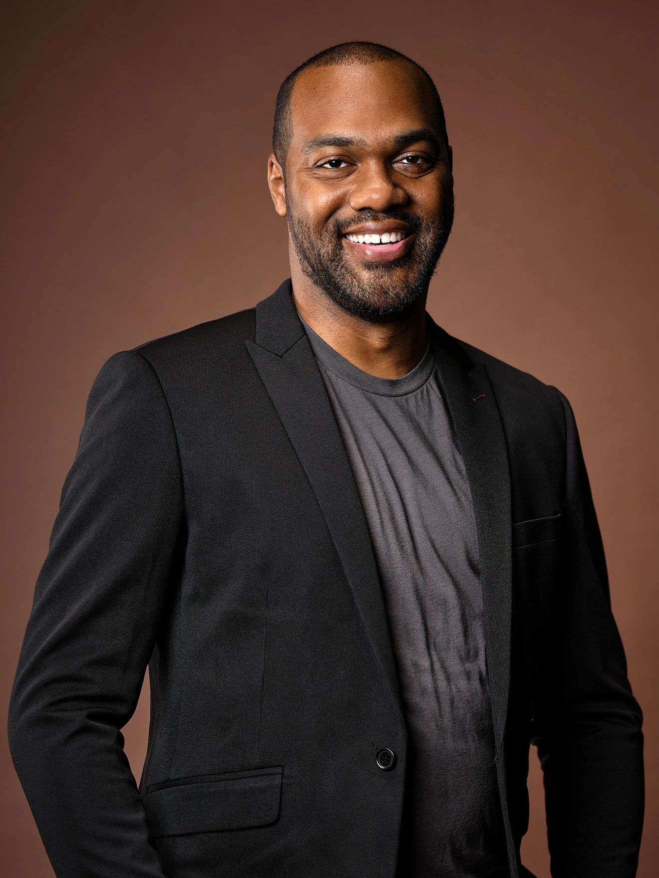 Smiling man in a black blazer and gray shirt against a brown background.