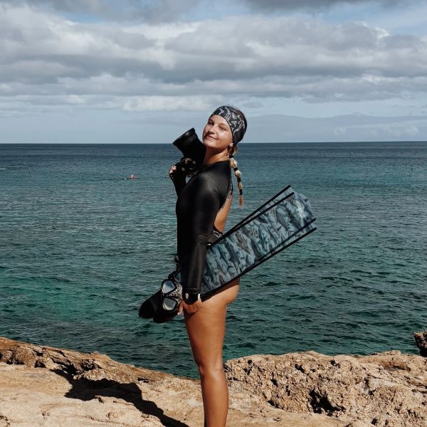 Woman in dive gear by the ocean on rocky shore.
