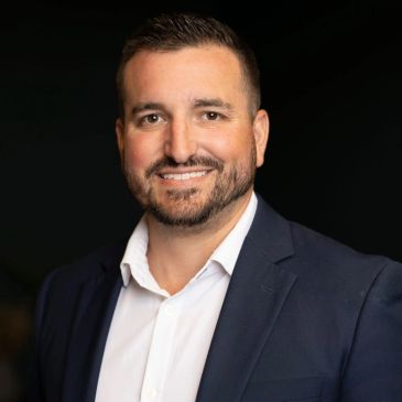 Professional man in a navy blazer and white shirt smiling against a dark background.