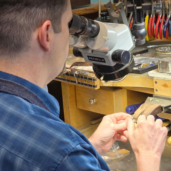 A man sitting at a bench looking through a microscope and working with his hands.