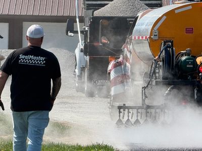 A worker in a SealMaster shirt oversees road paving with heavy machinery.