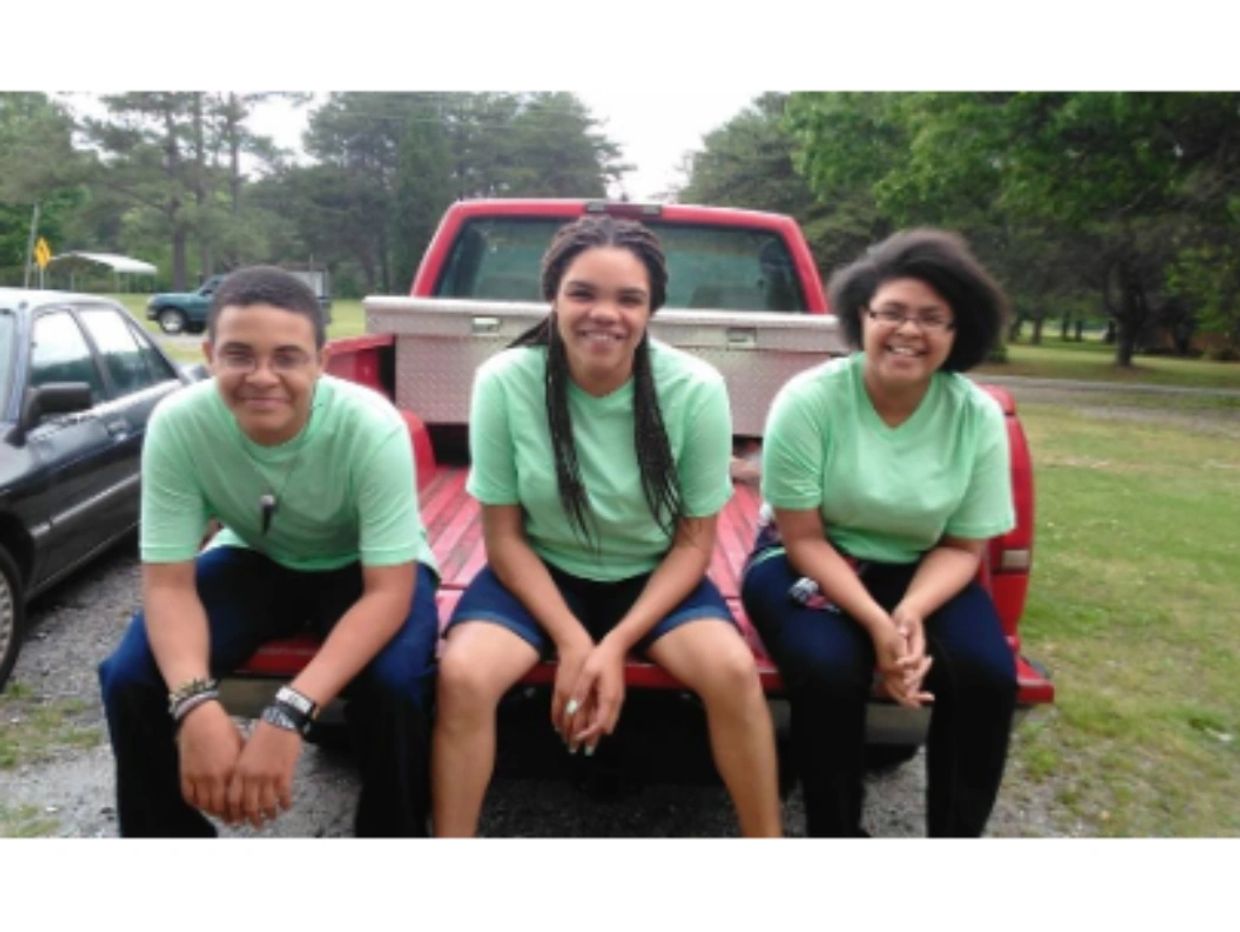 Three young people in green shirts sitting on a red pickup truck bed outdoors.