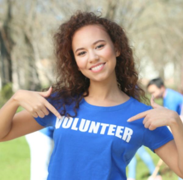 Smiling woman pointing to her blue "VOLUNTEER" t-shirt outdoors.