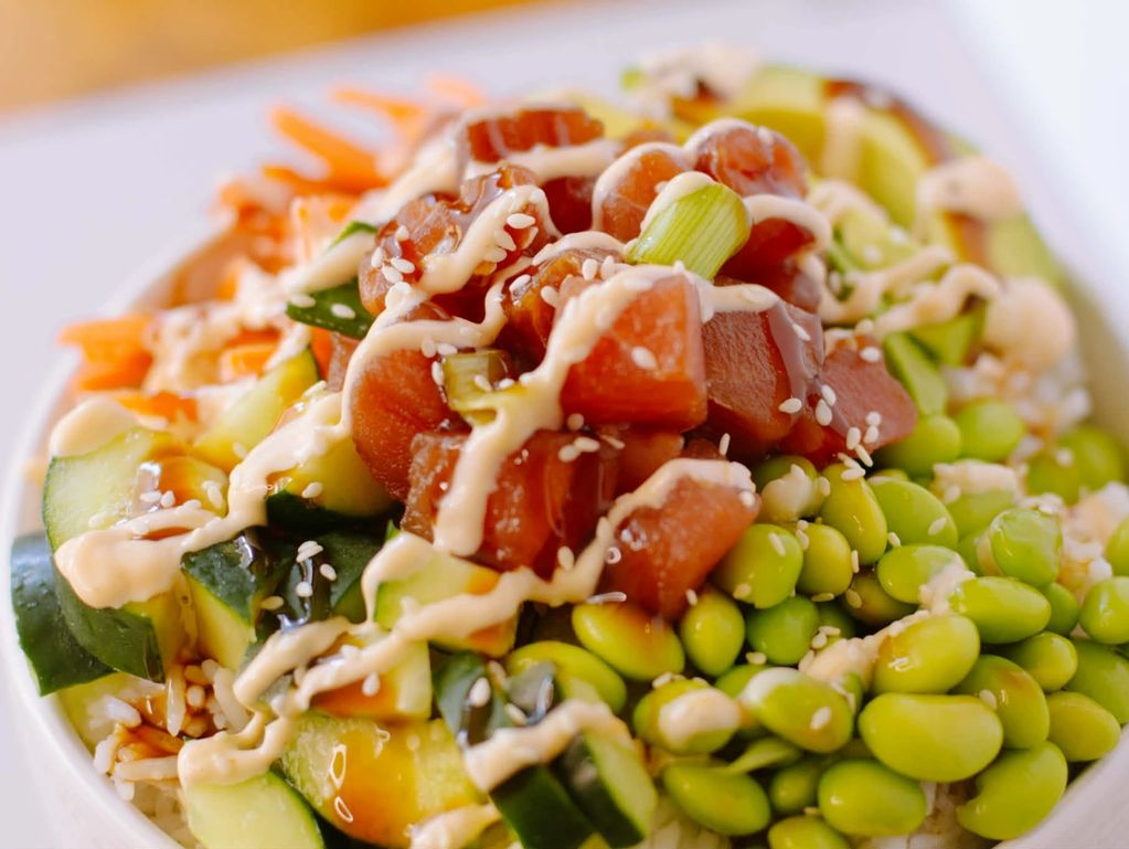 A colorful poke bowl with diced fish, edamame, cucumber, and drizzle of sauce.