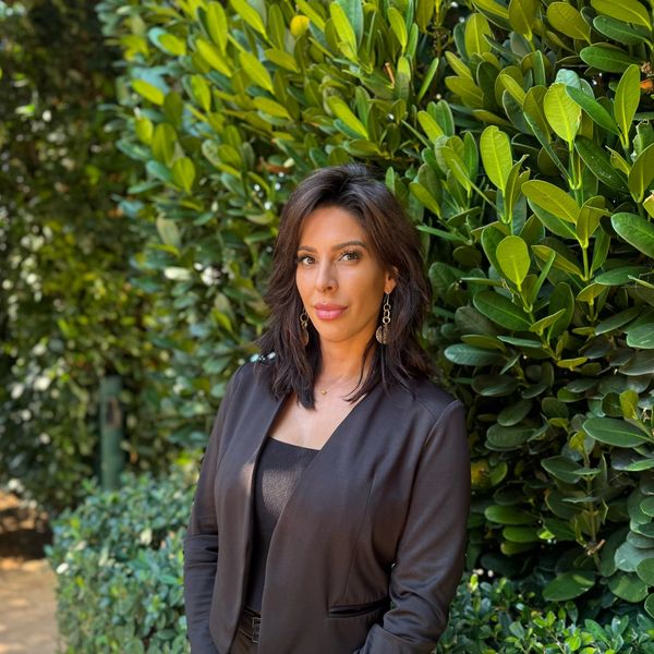 Woman in black blazer standing by lush green foliage.