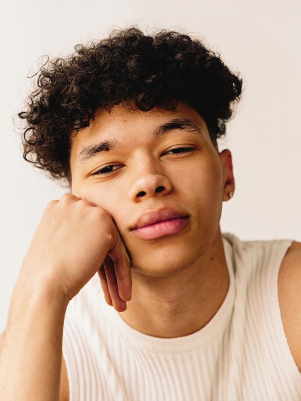 Young man with curly hair resting his face on his hand, wearing a white sleeveless shirt.