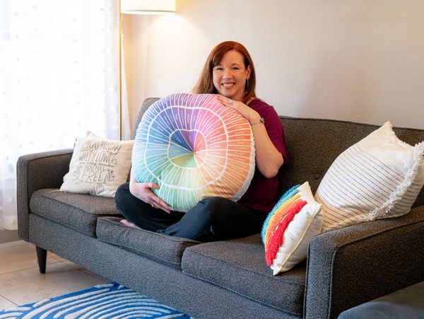 Smiling woman with red hair is sitting on a couch, holding a feelings-wheel pillow 