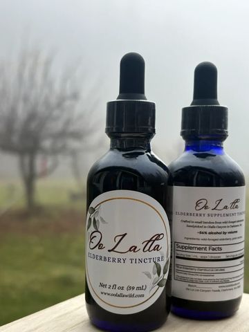 Two bottles of Oo La Lla Elderberry Tincture on a wooden surface with a foggy background.