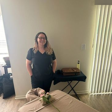 A woman in black scrubs stands in a softly lit room with a massage table and a flower.