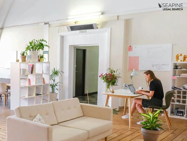 A woman working on a laptop in a bright, cozy office with plants and a white couch.