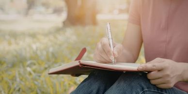 Person writing in a notebook outdoors on a sunny day.