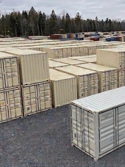 Rows of stacked shipping containers in an outdoor storage area near trees.