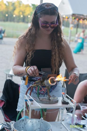 A woman creates glass art using a torch flame outdoors.