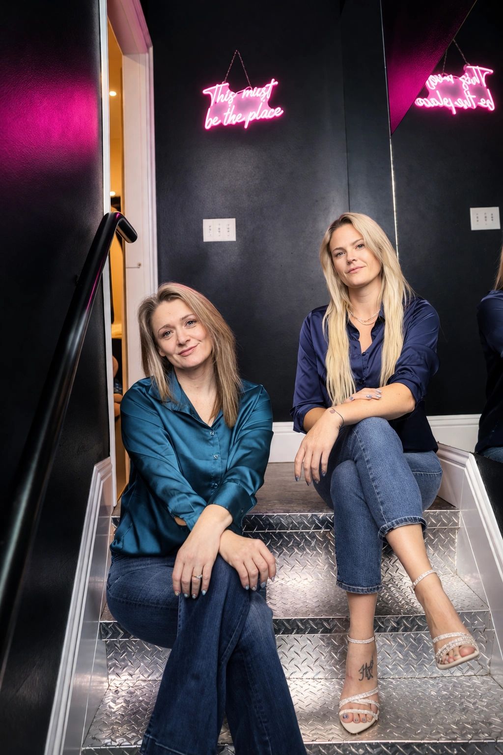 Two women sitting on metal stairs under pink neon sign.