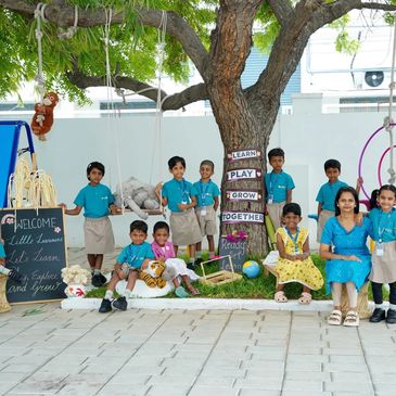 Group of children and teacher outdoors with welcome sign and tree.