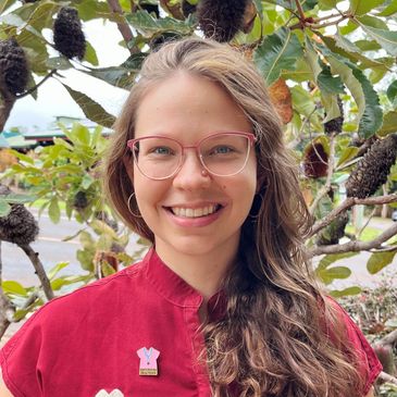 Smiling woman with glasses and long hair in a red shirt, standing outdoors with greenery.